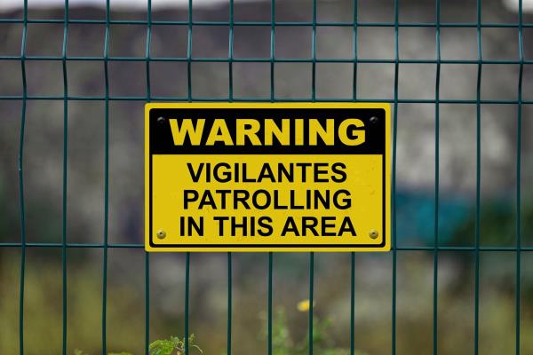 Red an white warning sign on a fence stating in "Warning - Vigilantes patrolling in this area" with a blank space underneath.