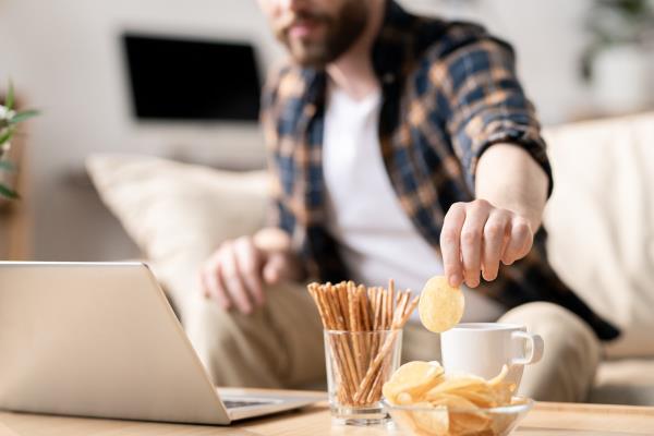 Young man taking potato chip out of glass bowl while sitting on sofa in front of laptop on table and havin<em></em>g snack